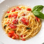 A white plate of Spaghetti al Pomodoro, topped with diced tomatoes, grated cheese, and a garnish of fresh basil leaves, set on a marble surface.