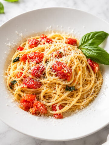 A white plate of Spaghetti al Pomodoro, topped with diced tomatoes, grated cheese, and a garnish of fresh basil leaves, set on a marble surface.
