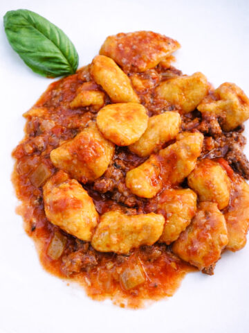 A plate of Gnocchi con Patate e Zucca con Ragu' alla Bolognese, topped with rich tomato meat sauce and garnished with a fresh basil leaf on the side.