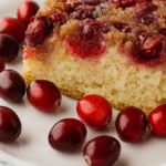 A close-up of a Cranberry Upside Down Cake slice on a white plate with a blue floral pattern, surrounded by whole fresh cranberries.