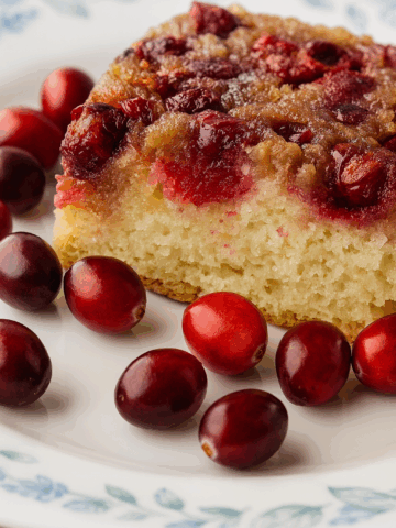 A close-up of a Cranberry Upside Down Cake slice on a white plate with a blue floral pattern, surrounded by whole fresh cranberries.