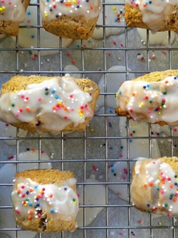 Mostaccioli-Christmas Spice Cookies, topped with rainbow sprinkles, are cooling on a wire rack. Drips of white icing and sprinkles add festive color to the surface below.
