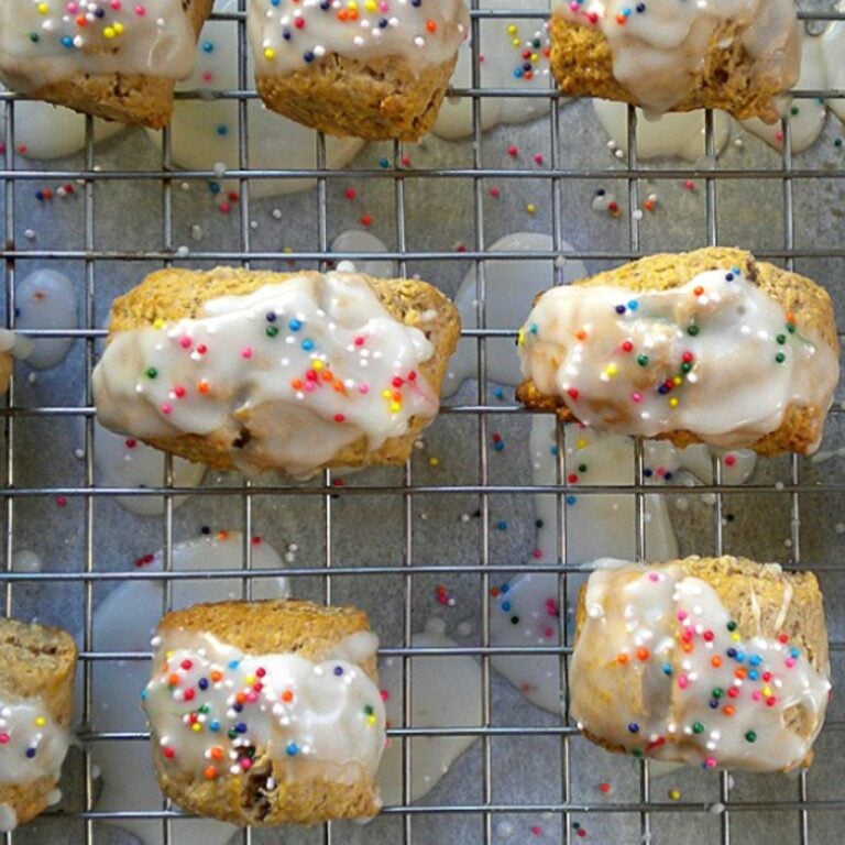 Mostaccioli-Christmas Spice Cookies, topped with rainbow sprinkles, are cooling on a wire rack. Drips of white icing and sprinkles add festive color to the surface below.