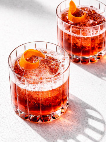 Two glasses of Negroni Sbagliato with ice cubes are garnished with orange peel twists. The cocktails sit on a light surface, illuminated by bright lighting that casts clear shadows.