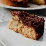 A close-up of a slice of crumbly, golden-brown Coffee Cake with a cinnamon and nut topping, served on a white plate. The rest of the coffee cake waits in the background on another plate.