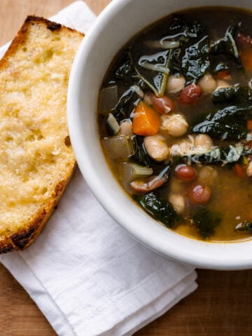 A bowl of Zuppa di Fagioli—Italian bean soup—filled with leafy greens, carrots, and beans sits next to a slice of toasted bread with melted cheese on a white napkin, all set on a wooden surface.