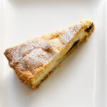A triangular slice of Crostata alla Ricotta and Chocolate Chips is dusted with powdered sugar and served on a white square plate, revealing tempting bits of fruit or filling inside the cake.
