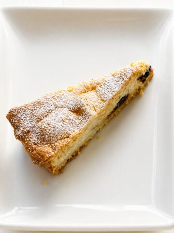 A triangular slice of Crostata alla Ricotta and Chocolate Chips is dusted with powdered sugar and served on a white square plate, revealing tempting bits of fruit or filling inside the cake.