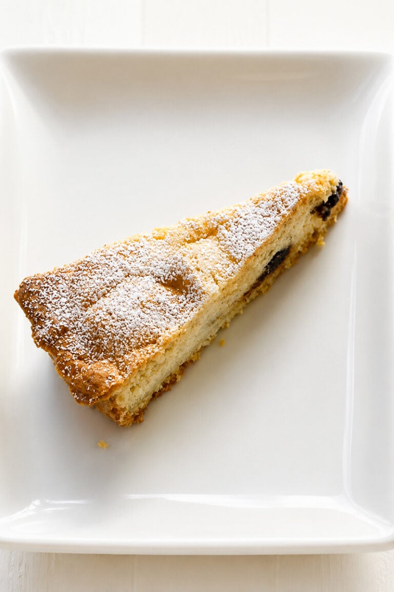 A triangular slice of Crostata alla Ricotta and Chocolate Chips is dusted with powdered sugar and served on a white square plate, revealing tempting bits of fruit or filling inside the cake.