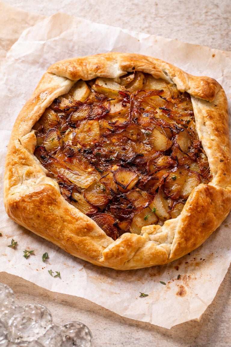 A rustic savory tart with potatoes, caramelized onion, and creamy feta in a golden, flaky crust sits on parchment paper, garnished with fresh thyme, atop a light stone surface.