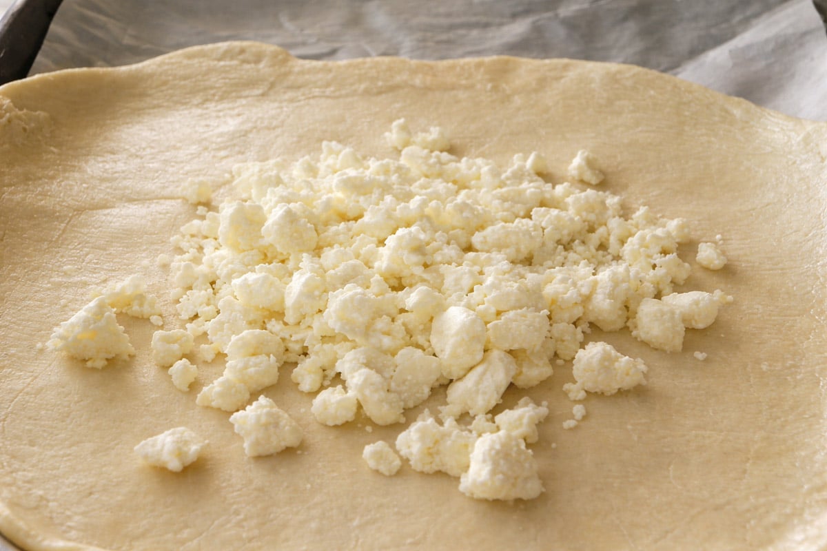 Crumbled feta cheese is spread on top of a rolled-out sheet of dough, ready to be folded or baked for a savory tart with potatoes, with a parchment-lined baking tray underneath.