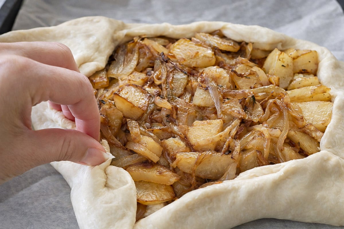 A close-up of a hand folding the edge of raw pastry dough over a filling of sautéed potatoes, onions, and creamy feta on parchment paper, preparing to bake a rustic savory tart with potatoes.
