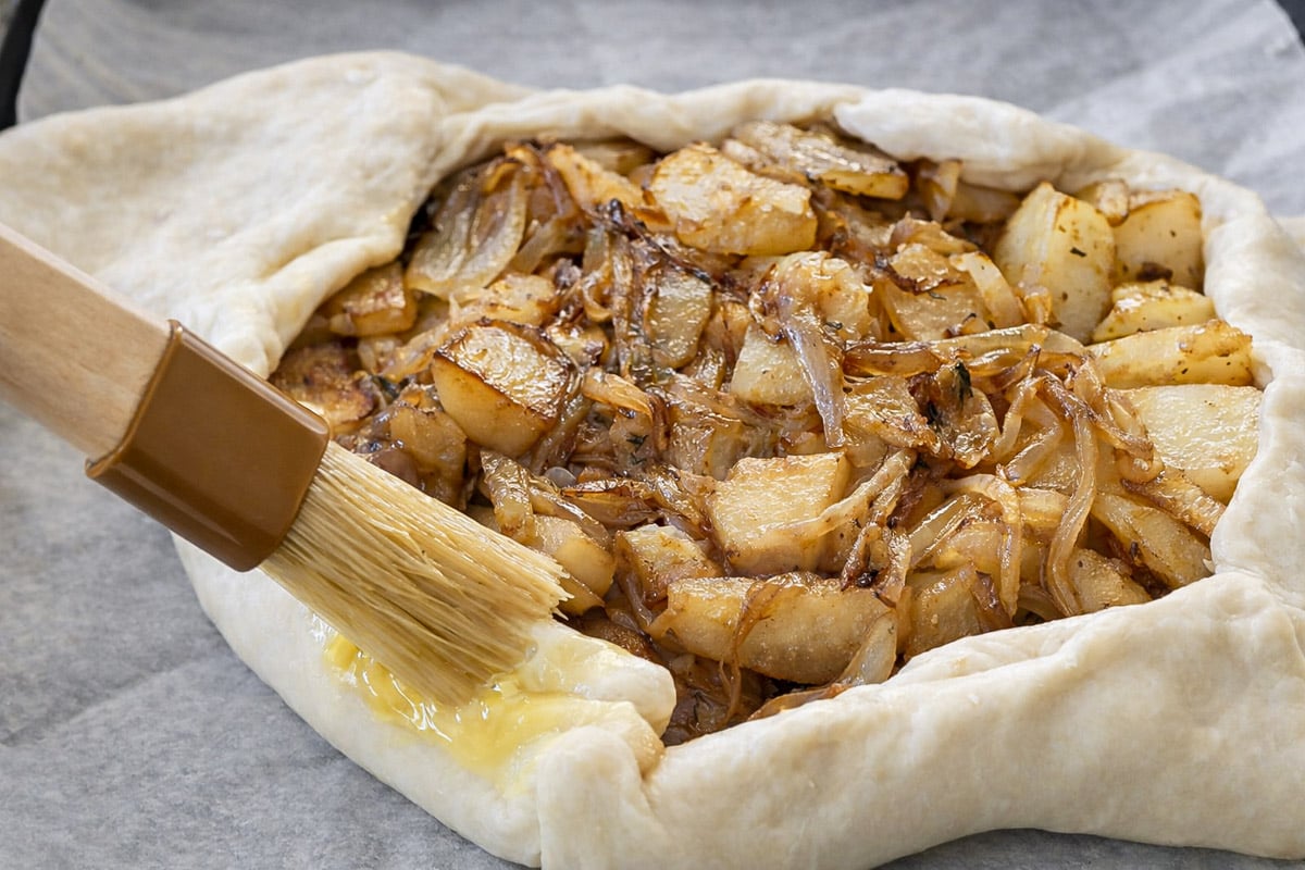 A pastry crust for a savory tart with potatoes is being brushed with an egg wash, encasing a filling of cooked potatoes, onions, and creamy feta on a sheet of parchment paper.