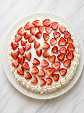 A round Traditional Italian Birthday Cake topped with a layer of whipped cream and decorated with sliced fresh strawberries, displayed on a white plate on a marble surface.