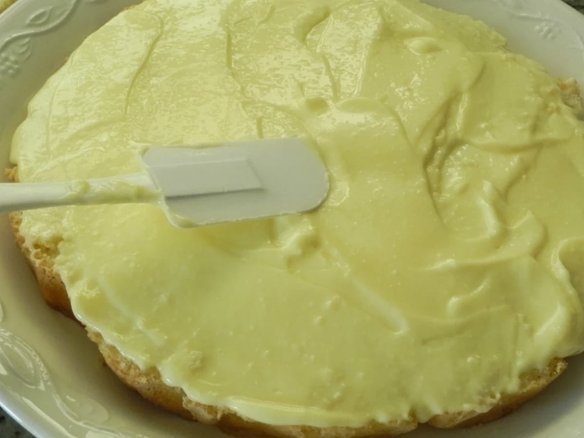 A close-up of a Traditional Italian Birthday Cake layer being spread with creamy yellow frosting using a white spatula on a white plate.