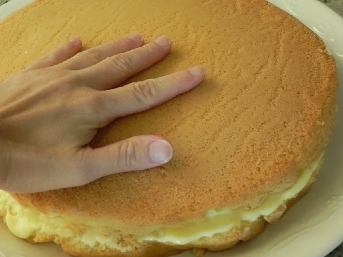 A hand pressing gently on top of a round Traditional Italian Birthday Cake filled with cream, placed on a white plate.