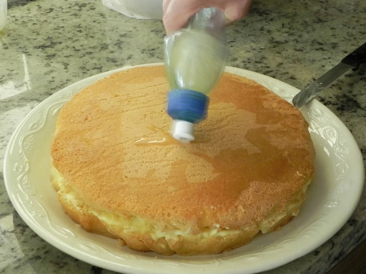 A hand squeezes syrup from a plastic bottle onto a round Traditional Italian Birthday Cake placed on a white plate, set on a granite countertop.