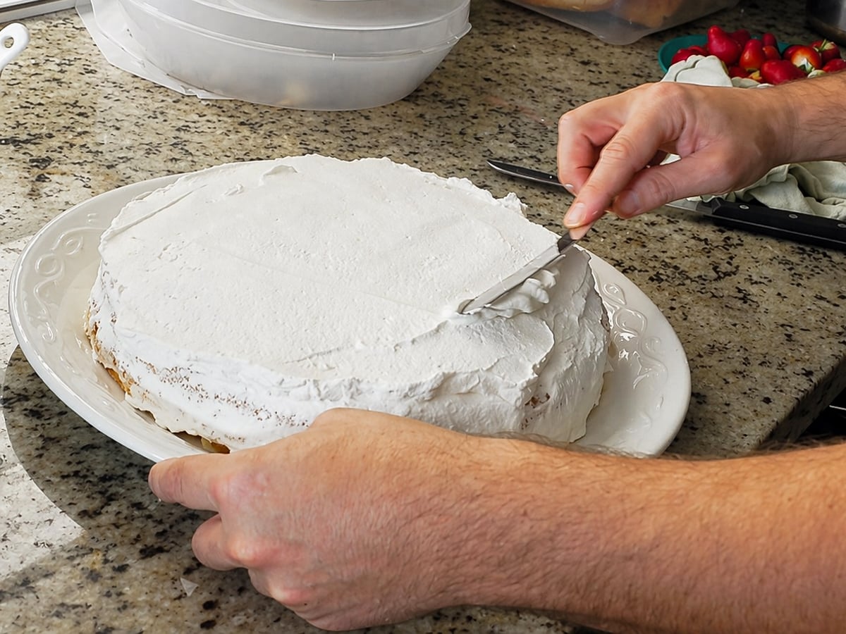 A person spreads white frosting on a round Traditional Italian Birthday Cake with a spatula, working on a granite countertop. Strawberries and a container are visible in the background.