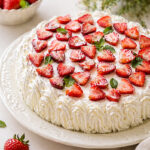 A Traditional Italian Birthday Cake, this round delight is topped with sliced strawberries and mint leaves, decorated with piped whipped cream. Fresh strawberries and a bowl of berries sit nearby, framed by a bouquet of small white flowers in the background.