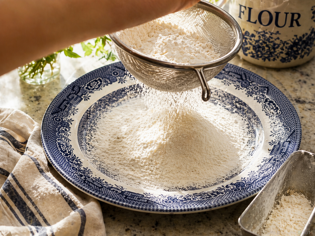 A hand sifting flour through a metal sieve onto a blue and white patterned plate, preparing ingredients for a Traditional Italian Birthday Cake, with a flour container and striped kitchen towel nearby on a sunlit kitchen counter.