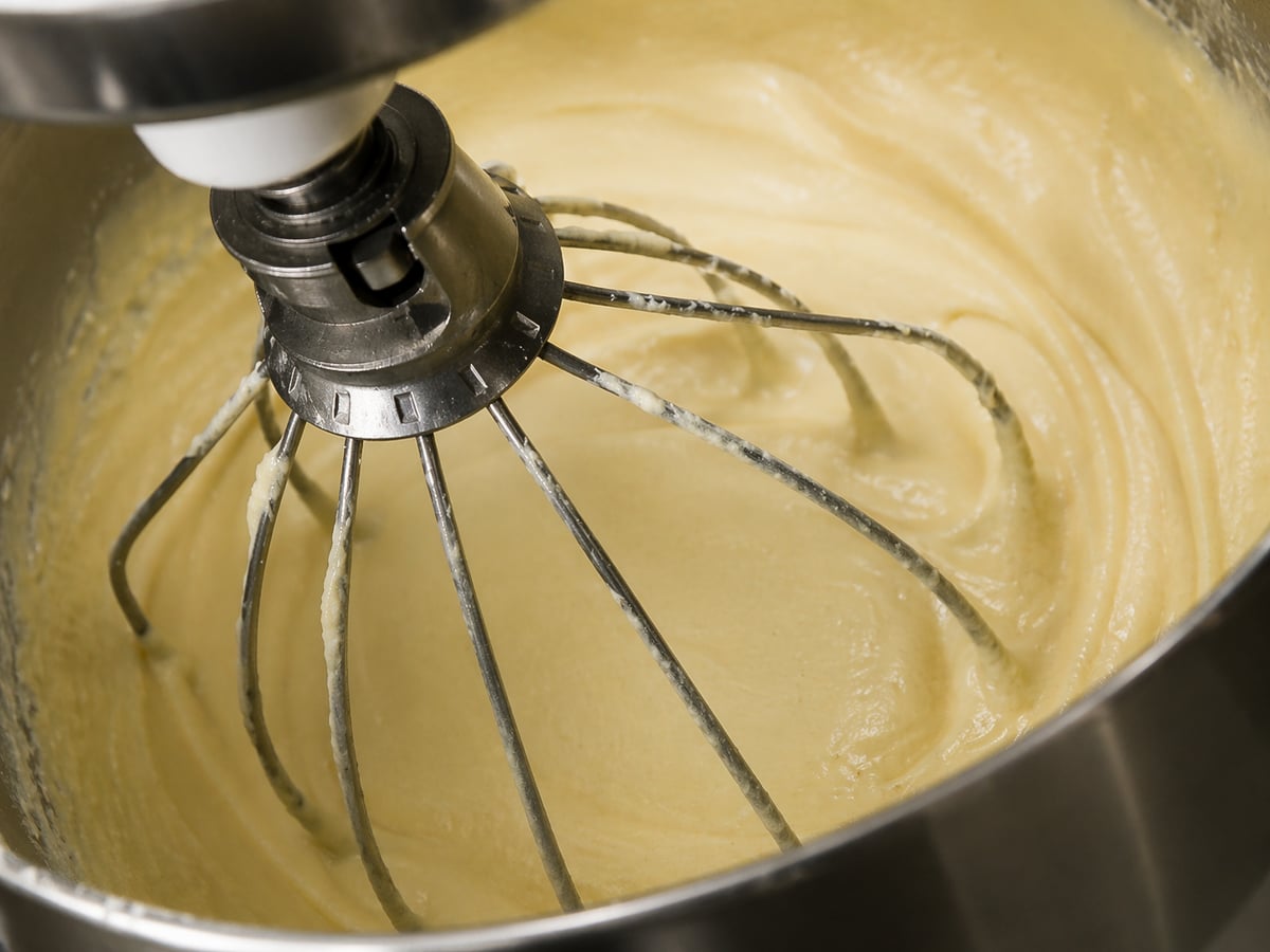 Close-up of a stand mixer with a whisk attachment blending smooth, creamy cake batter for a traditional Italian birthday cake in a metal mixing bowl.