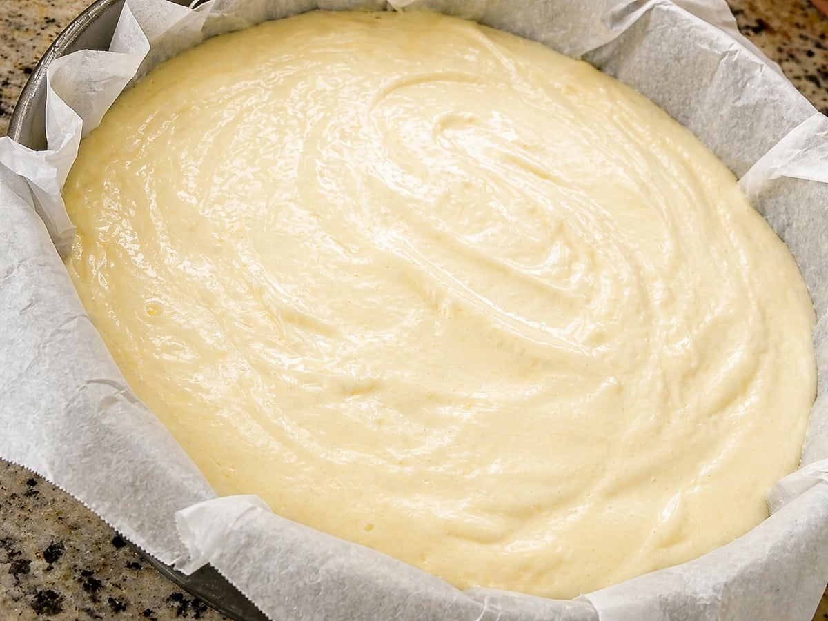 Cake batter for a Traditional Italian Birthday Cake is spread evenly in a round baking pan lined with parchment paper, ready to be baked. The pan rests on a speckled kitchen countertop.