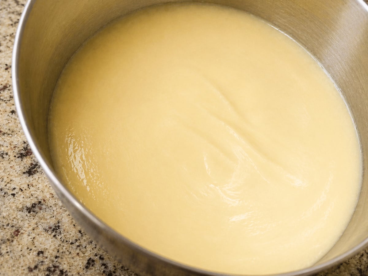 A metal bowl filled with smooth, creamy, light yellow batter or dough—perfect for making a Traditional Italian Birthday Cake—sits on a speckled countertop.