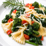 Plate of Farfalle con Broccoli Rapini with sautéed broccolini, chopped tomatoes, and grated cheese, garnished with a sprig of rosemary on a white plate.