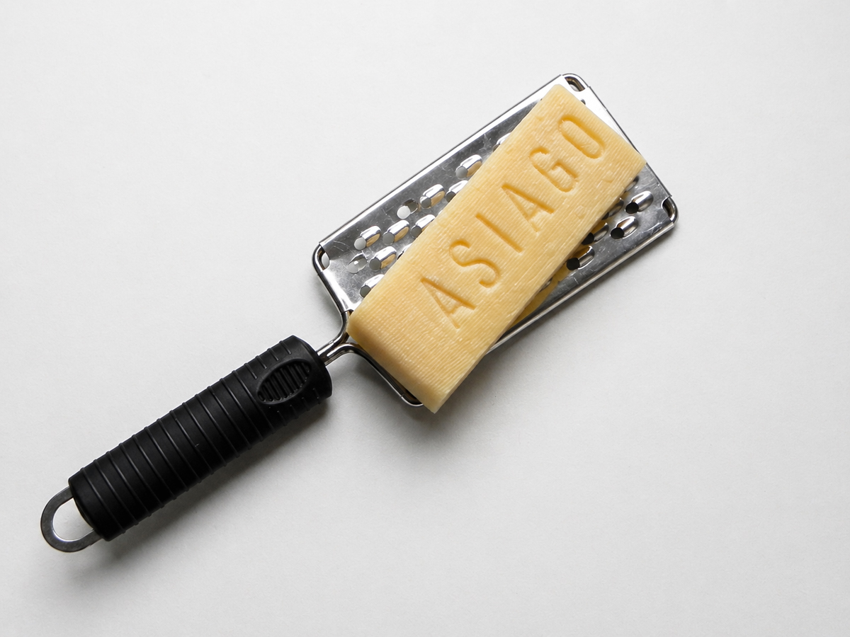 A block of Asiago cheese, perfect for topping Farfalle con Broccoli Rapini, is placed on a metal cheese grater with a black handle, on a white background. The word ASIAGO is engraved on the cheese block.