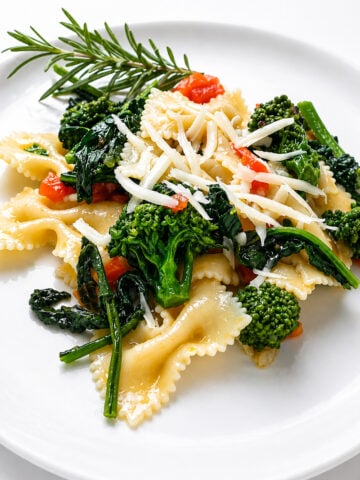 A plate of Farfalle con Broccoli Rapini features bow-tie pasta mixed with broccoli, sautéed greens, red bell peppers, and topped with shredded cheese, garnished with a sprig of rosemary.