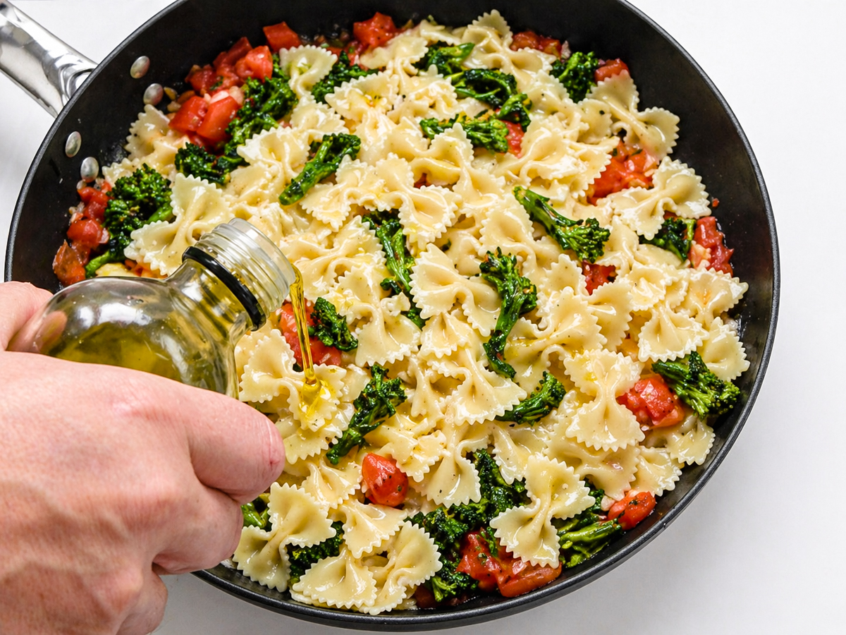 A hand pours olive oil over Farfalle con Broccoli Rapini, tossing bowtie pasta with tomatoes and broccoli in a skillet on a white surface.