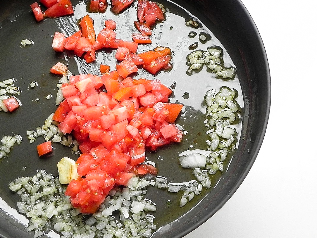 Chopped tomatoes, onions, and garlic sizzle in olive oil in a black pan-classic flavors perfect as a base for Farfalle con Broccoli Rapini-set against a clean white background.
