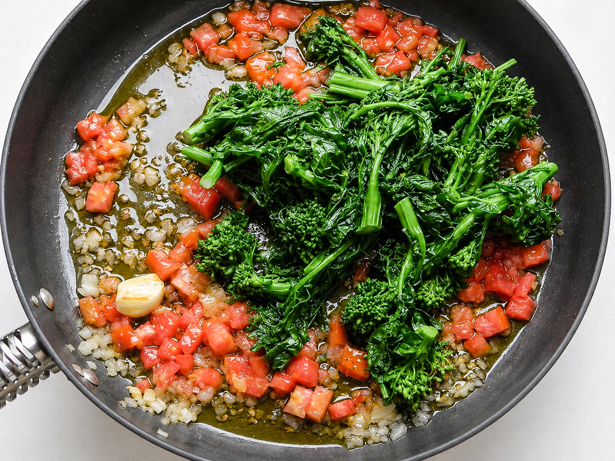 A skillet filled with sautéed diced onions, chopped tomatoes, rapini (broccoli rabe), garlic cloves, and olive oil, all cooking together on a stovetop—the perfect base for Farfalle con Broccoli Rapini.