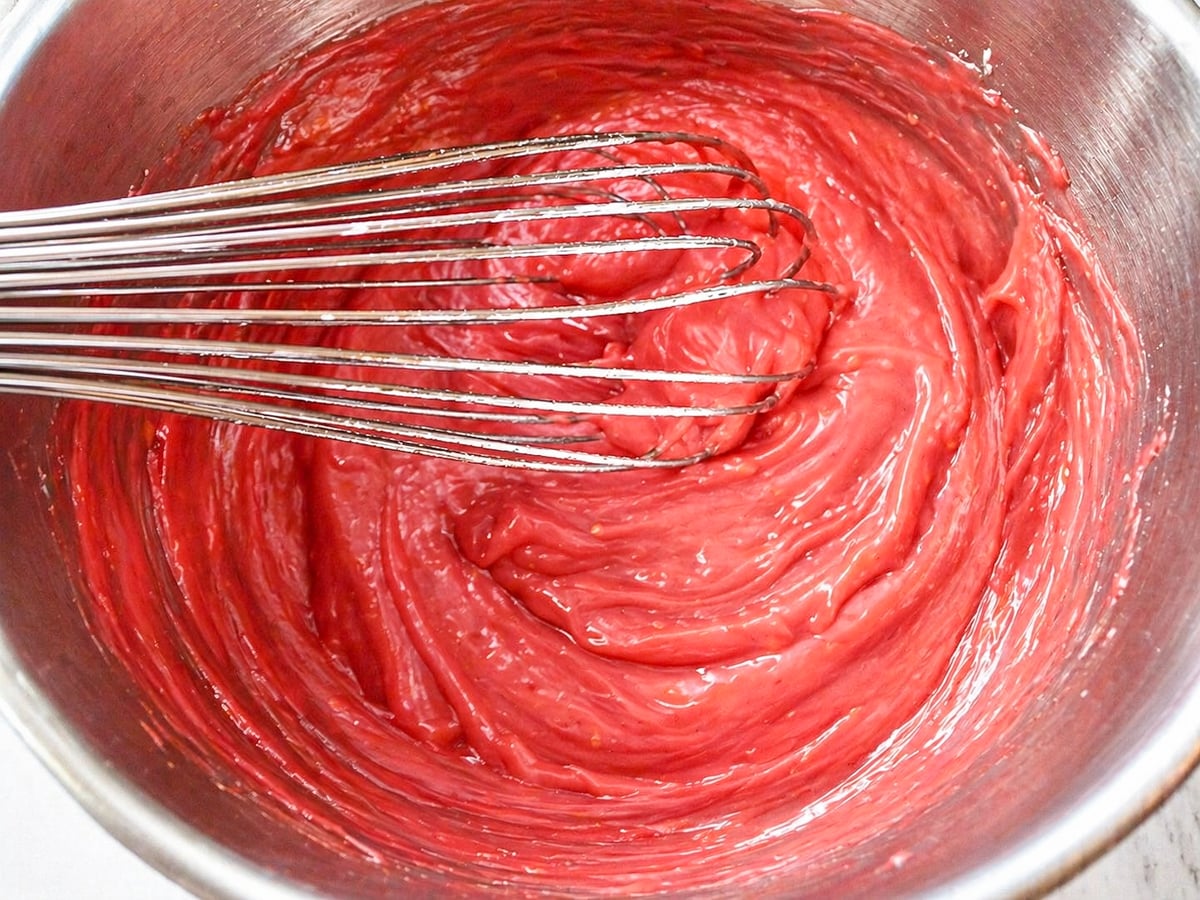 A metal whisk rests in a mixing bowl filled with smooth, thick red batter, likely the first step in preparing a delicious Pomegranate-Mixed Berry Pie or similar dessert.