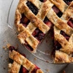 A lattice-topped Pomegranate-Mixed Berry Pie in a glass dish with a golden, flaky crust. One slice sits on the table nearby, revealing the colorful berry and pomegranate filling. A pie server rests next to the dessert.