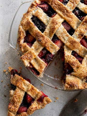 A lattice-topped Pomegranate-Mixed Berry Pie in a glass dish with a golden, flaky crust. One slice sits on the table nearby, revealing the colorful berry and pomegranate filling. A pie server rests next to the dessert.
