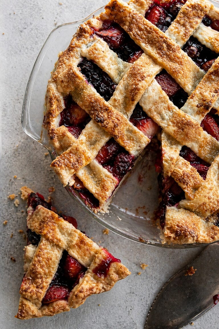 A lattice-topped Pomegranate-Mixed Berry Pie in a glass dish with a golden, flaky crust. One slice sits on the table nearby, revealing the colorful berry and pomegranate filling. A pie server rests next to the dessert.
