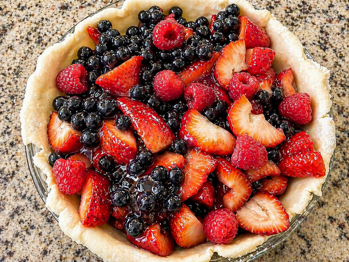 An unbaked Pomegranate-Mixed Berry Pie crust filled with fresh sliced strawberries, whole raspberries, and blueberries sits on a speckled countertop.