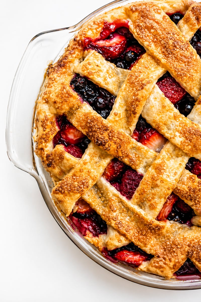 A lattice-topped Mixed Berry Pie in a glass dish with a golden, flaky crust. One slice sits on the table nearby, revealing the colorful berry and pomegranate filling. A pie server rests next to the dessert.
