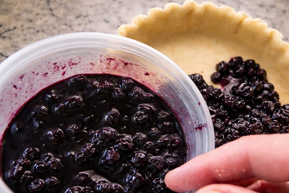A close-up of a hand filling a pie crust with glossy, dark blueberry pie filling on a kitchen countertop—perfect for your next blueberry crostata recipe.