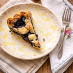 A slice of blueberry crostata, inspired by a classic blueberry crostata recipe, sits on a decorative plate with a fork on a cloth napkin. Two lemon wedges rest in the background on a wooden table.