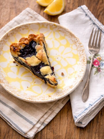 A slice of blueberry crostata, inspired by a classic blueberry crostata recipe, sits on a decorative plate with a fork on a cloth napkin. Two lemon wedges rest in the background on a wooden table.