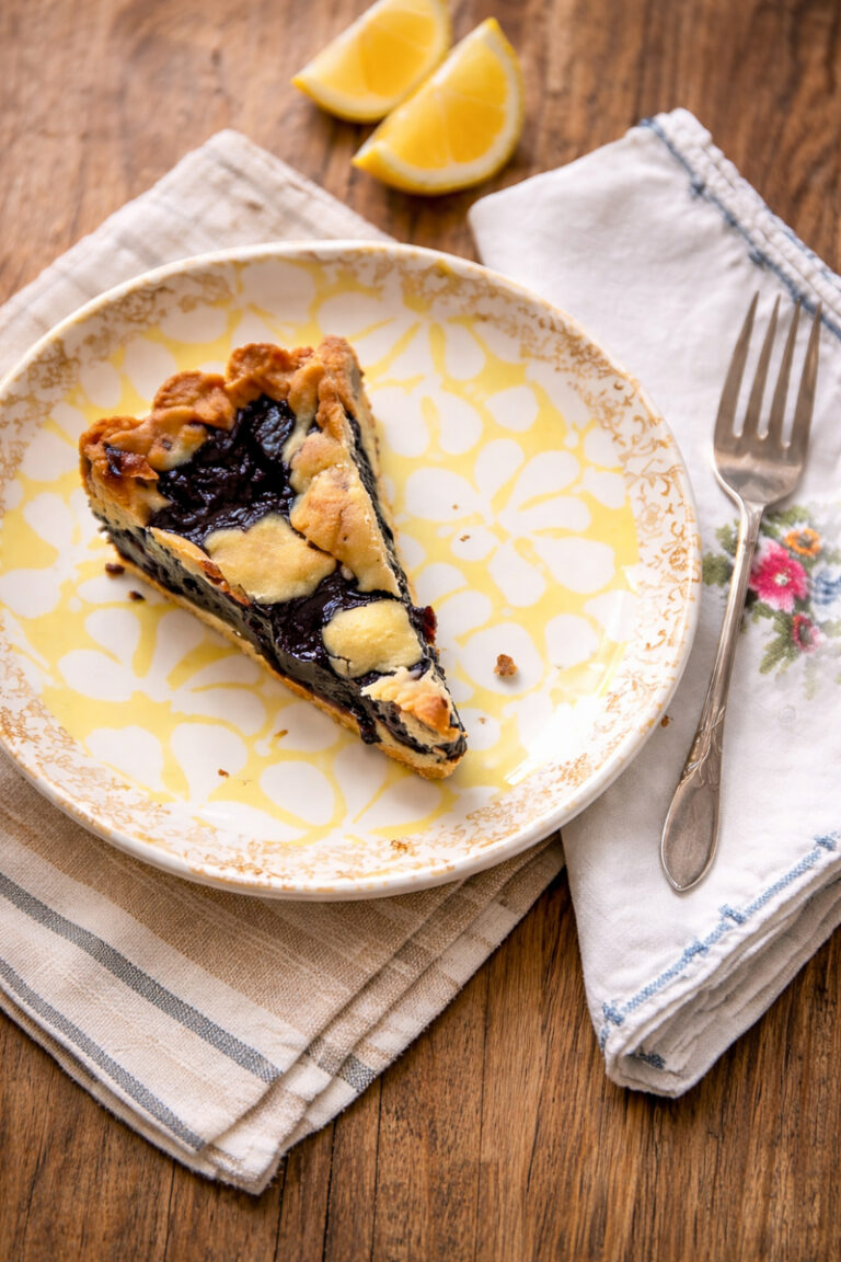 A slice of blueberry crostata, inspired by a classic blueberry crostata recipe, sits on a decorative plate with a fork on a cloth napkin. Two lemon wedges rest in the background on a wooden table.