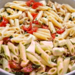 A close-up of a bowl filled with Penne with Mozzarella, cherry tomatoes, fresh basil, chopped herbs, and walnuts, all mixed together.