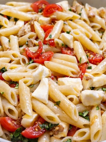 A close-up of a bowl filled with Penne with Mozzarella, cherry tomatoes, fresh basil, chopped herbs, and walnuts, all mixed together.