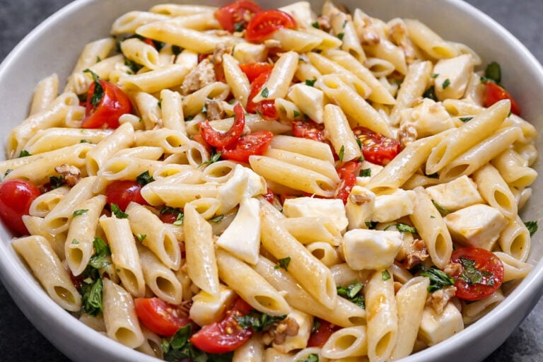 A close-up of a bowl filled with Penne with Mozzarella, cherry tomatoes, fresh basil, chopped herbs, and walnuts, all mixed together.