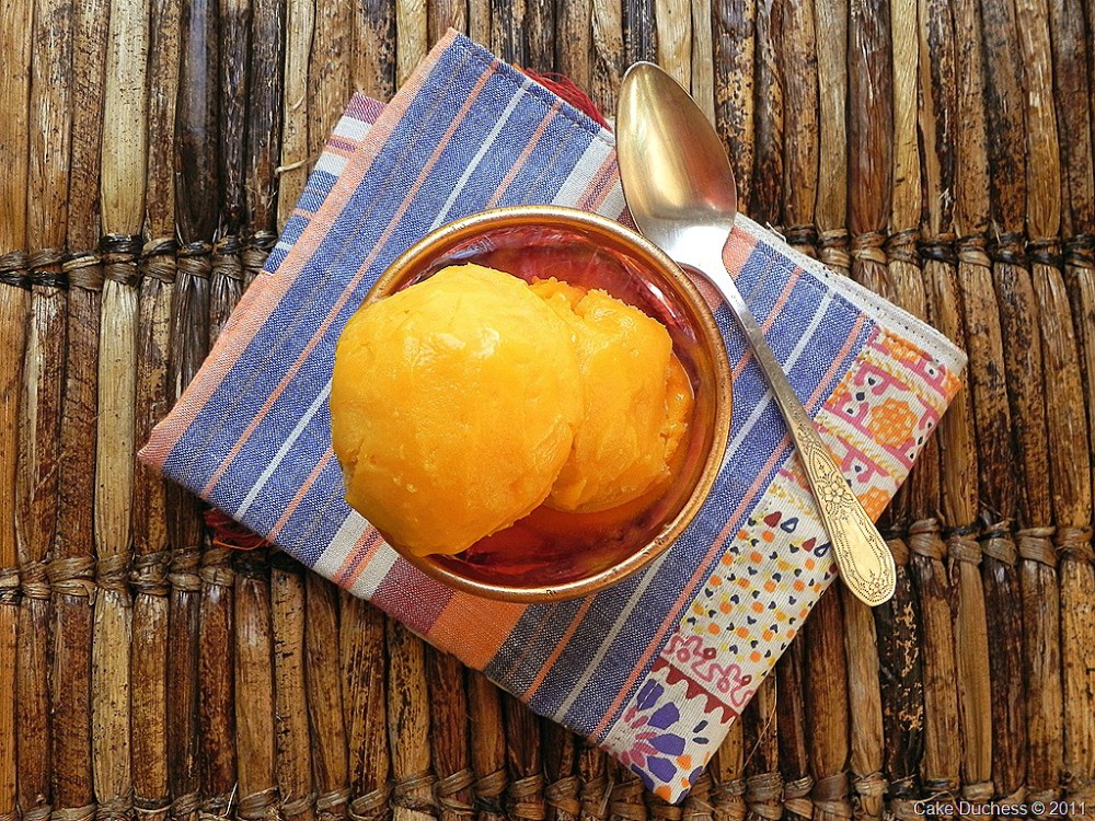 A bowl of Mango Sorbet with two scoops is placed on a colorful striped napkin, next to a spoon, on a woven surface.