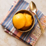 A bowl of creamy, bright yellow mango sorbet sits on two folded, patterned cloth napkins, with a silver spoon resting beside it on a light-colored surface.