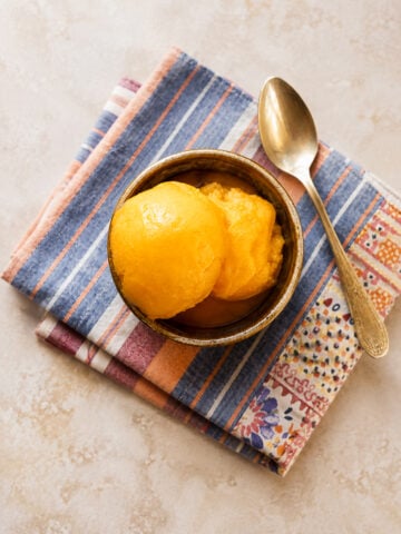 A bowl of creamy, bright yellow mango sorbet sits on two folded, patterned cloth napkins, with a silver spoon resting beside it on a light-colored surface.
