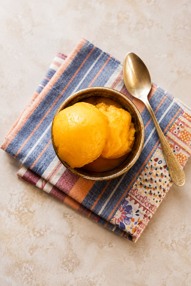 A bowl of creamy, bright yellow mango sorbet sits on two folded, patterned cloth napkins, with a silver spoon resting beside it on a light-colored surface.